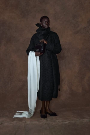 Model Athiec Geng wearing a dark textured coat and draped white fringed scarf against a brown textured backdrop.