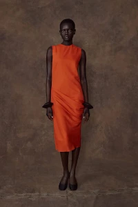 Model Athiec Geng wears an orange sleeveless midi dress and sculptural black cuffs in front of a textured brown backdrop.