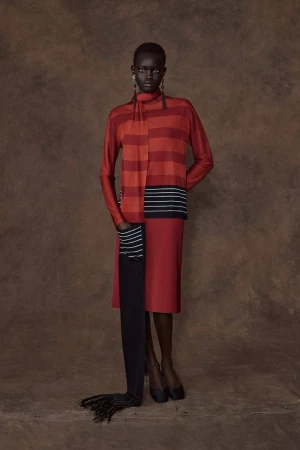 Model Athiec Geng standing in a red and orange striped knit top, matching skirt, and floor-length black scarf against a brown backdrop.
