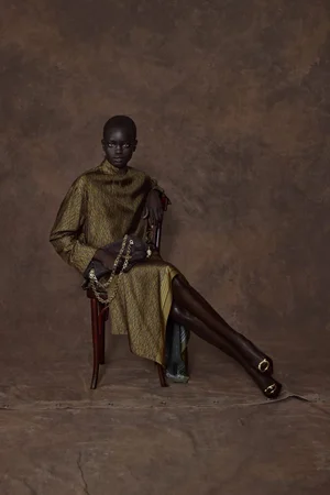 Model Athiec Geng seated on a wooden chair wearing a metallic gold jacquard dress for Ferragamo Pre-Fall 2026.