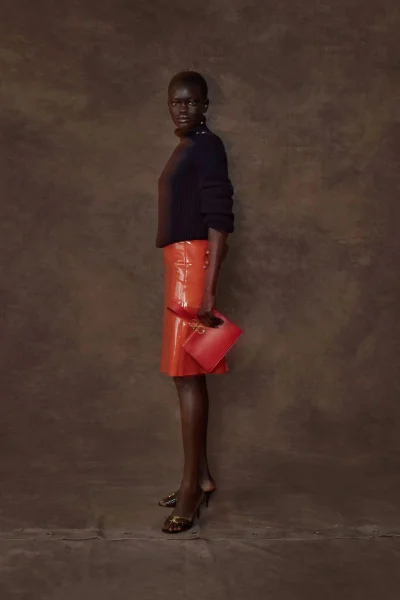 Athiec Geng poses in a dark knit sweater, glossy orange pencil skirt, and heels holding a red clutch against a textured brown backdrop.