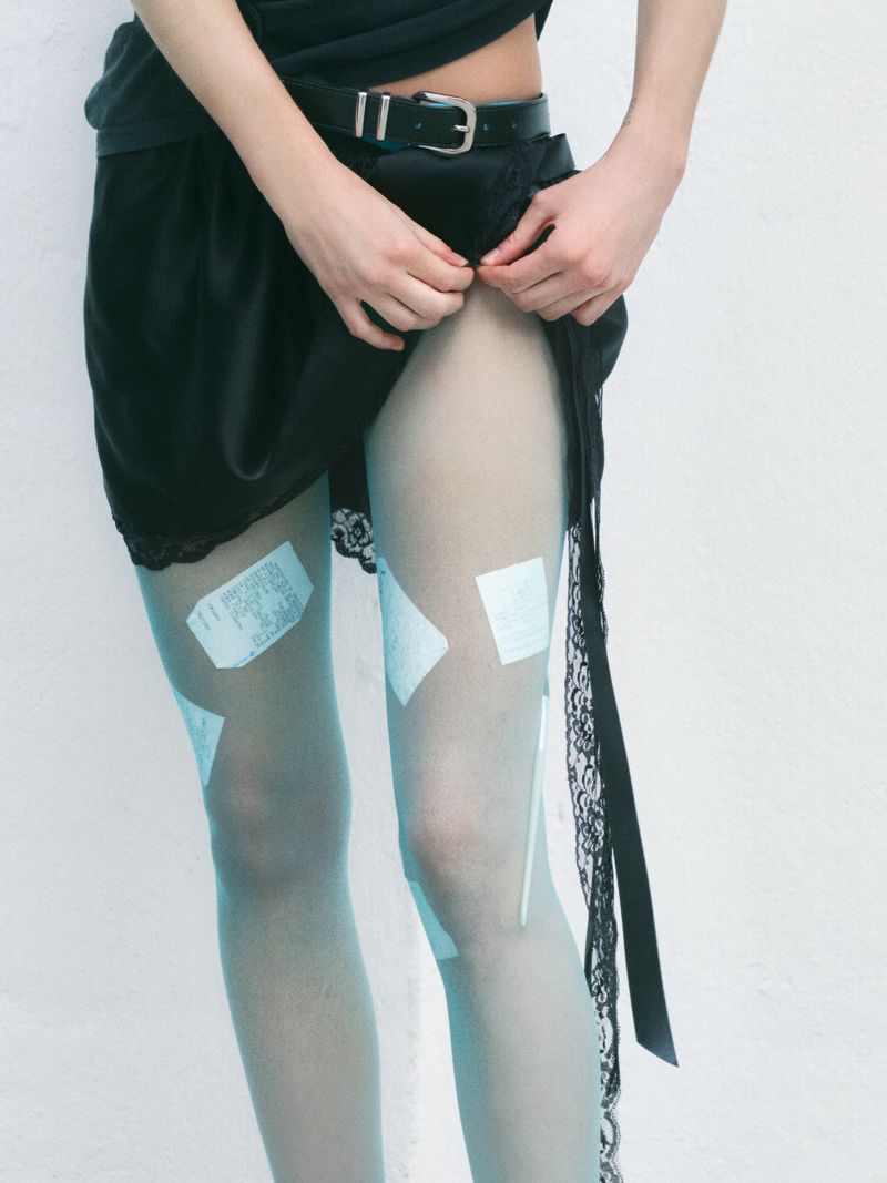 Close-up studio shot of a slender woman wearing a black top and skirt, with sheer blue tights and white labels taped to her legs.