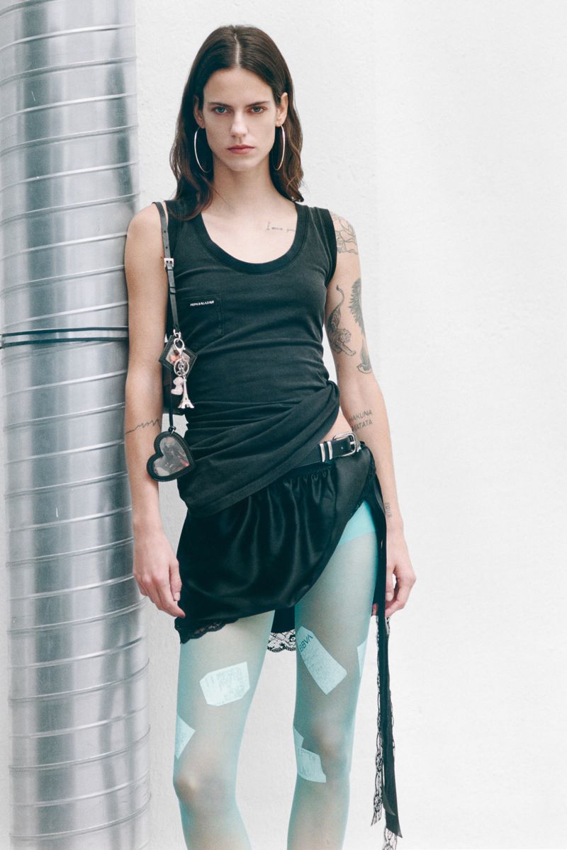 Fashionable woman in black tank top and skirt, posing against industrial background. She wears light blue tights with patches.