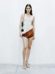 Model Miriam Sanchez standing against a white wall wearing an off-white tank top with feather trim, rust shorts, and a tiara.
