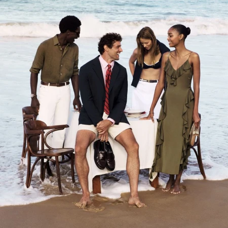 Four people gather around a table on a beach, two men and two women are in a relaxed setting with ocean waves in the background.
