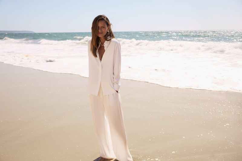A woman in a white button-down shirt and pants stands on a sandy beach with the ocean and waves in the background on a sunny day.