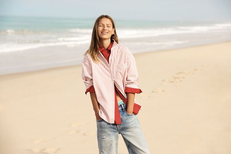 Adult woman in an unbuttoned striped shirt stands on a sandy beach with light-blue ocean and sky in the background on a sunny day.