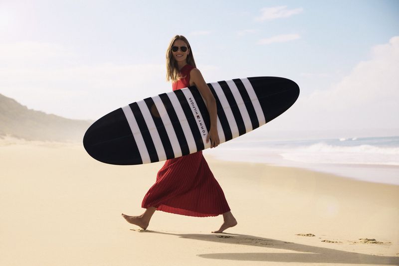 Woman in red dress carries a striped surfboard on a sandy beach under a bright sky, showcasing a blend of fashion and surf lifestyle.