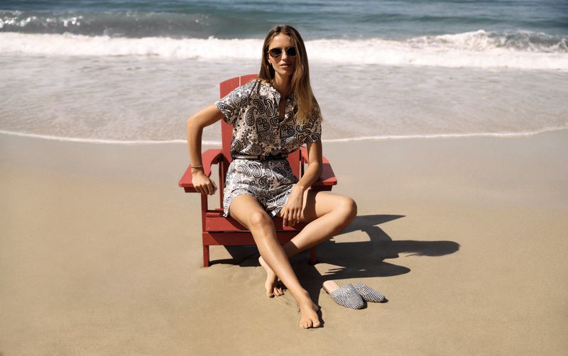 Woman in patterned summer outfit sits in red Adirondack chair on sandy beach with ocean waves, sunlit scene, casual fashion.