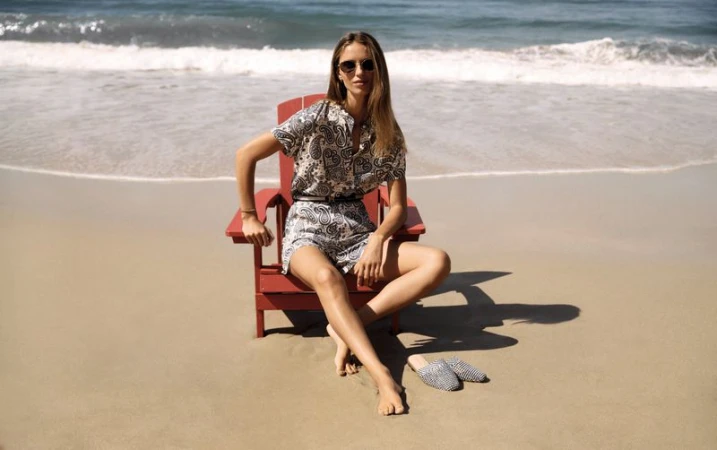 Woman in patterned summer outfit sits in red Adirondack chair on sandy beach with ocean waves, sunlit scene, casual fashion.