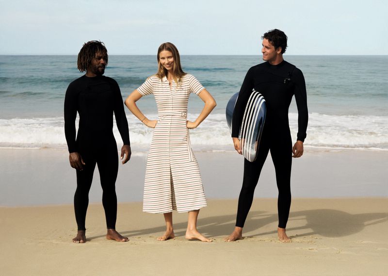 Three people, two men in wetsuits and a woman in a striped dress, pose on a sandy beach with the ocean in the background.