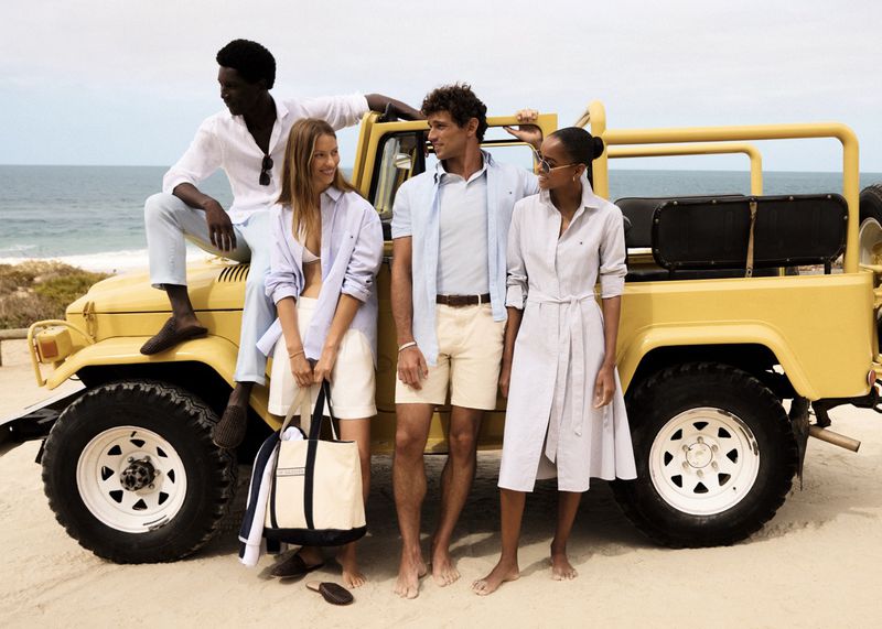 Group of four fashionably dressed people in light-colored clothing posing near a yellow jeep on a sandy beach with an ocean backdrop.