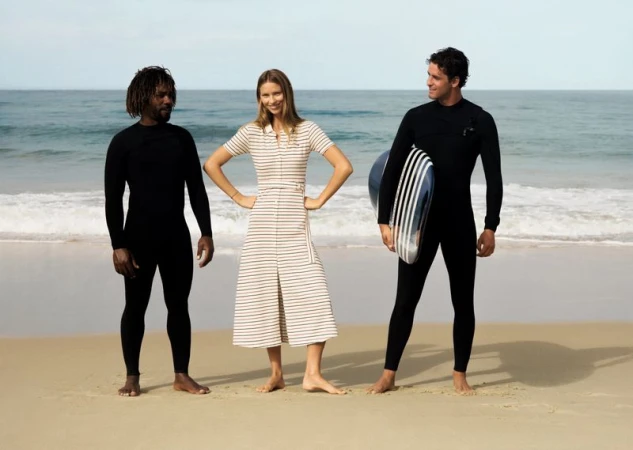 Three models standing barefoot on a sandy beach before ocean waves, two men in black wetsuits and a woman in a striped dress.