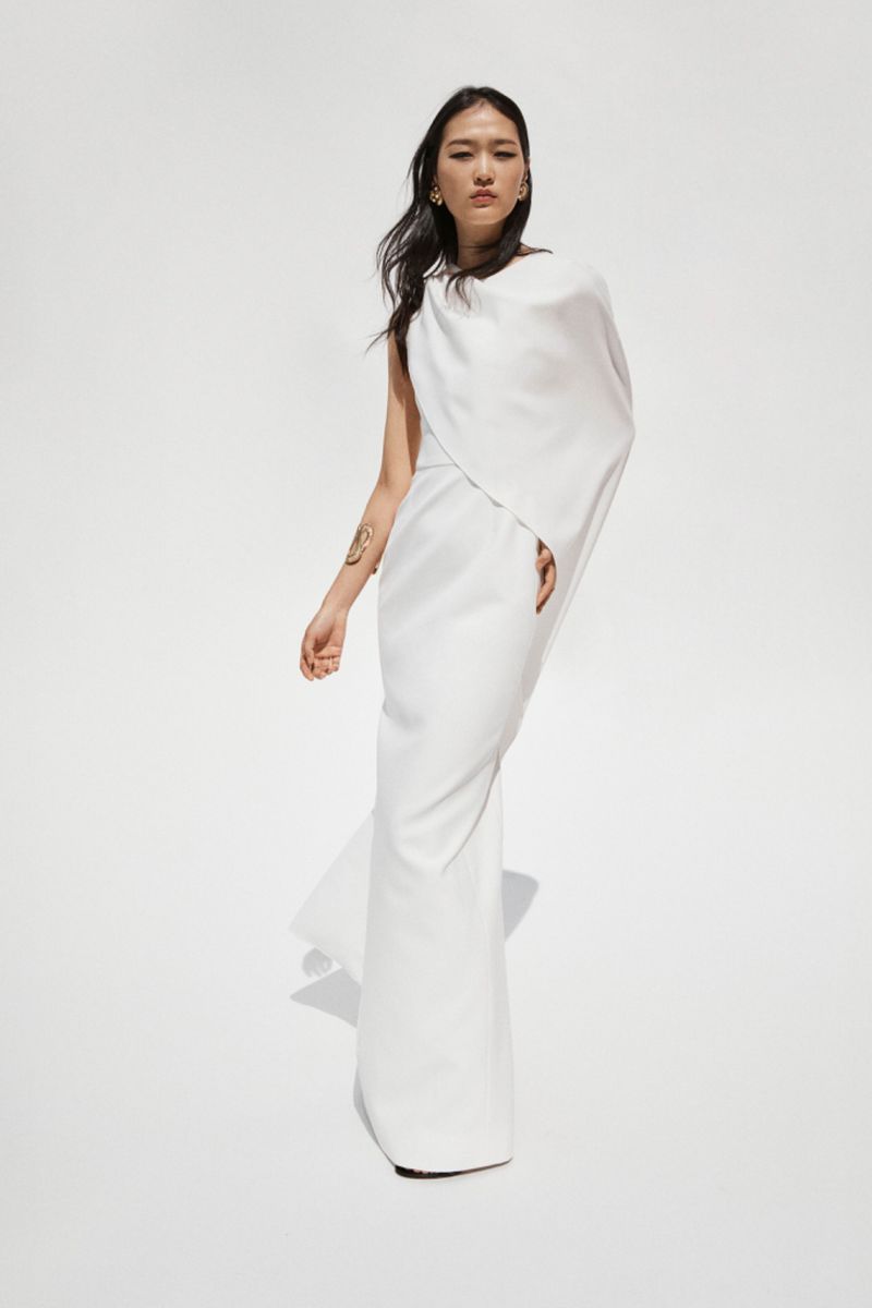 Elegant Asian woman in a white gown stands in a bright photo studio, showcasing modern fashion with simple draping and minimal jewelry.