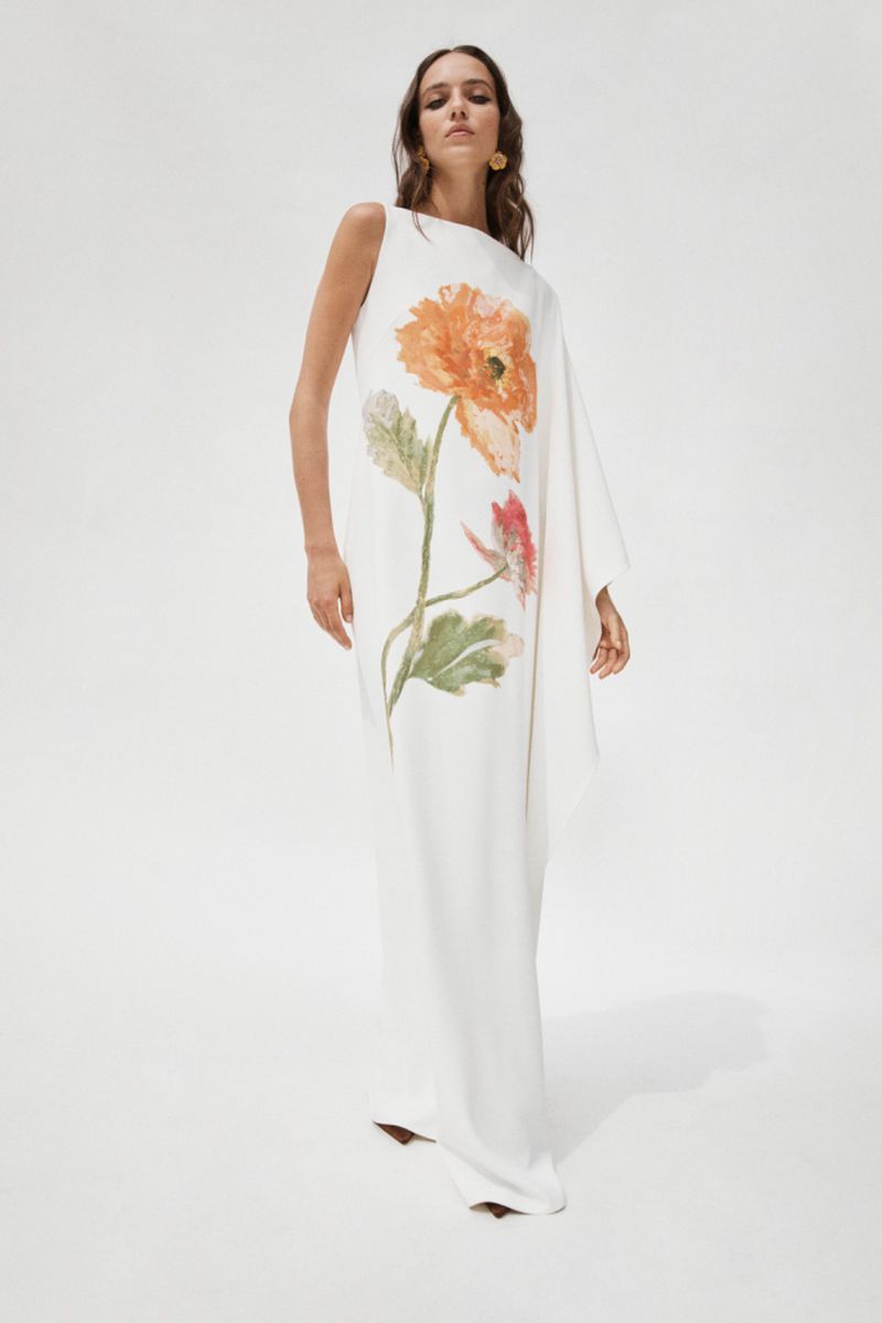 Elegant woman in a white, floor-length dress with floral design, posing in a bright studio. Fashion editorial shot.