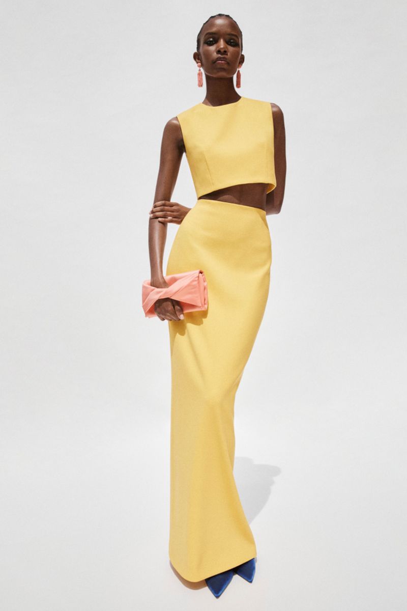 A woman poses in a yellow two-piece dress with a sleeveless crop top and long fitted skirt against a white backdrop, holding a clutch.