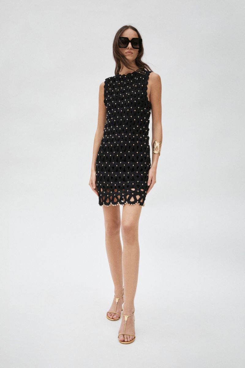 A slender woman poses in a sleeveless black mini-dress with embellishments, paired with brown sandals and sunglasses, against a white backdrop.