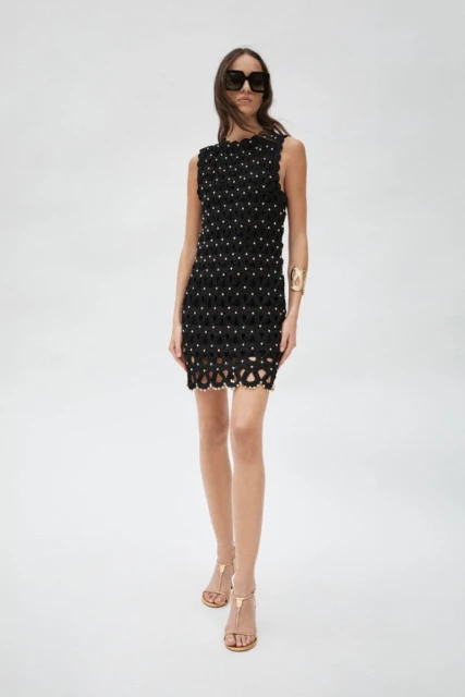 A slender woman poses in a sleeveless black mini-dress with embellishments, paired with brown sandals and sunglasses, against a white backdrop.