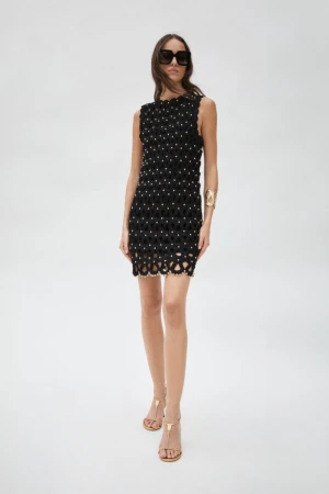 A slender woman poses in a sleeveless black mini-dress with embellishments, paired with brown sandals and sunglasses, against a white backdrop.