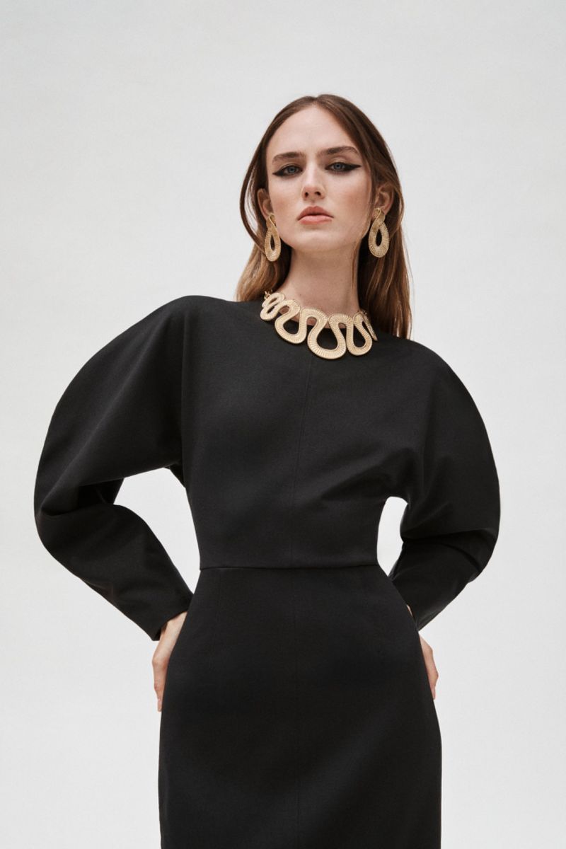 Studio shot of a model in a black midi dress with statement gold jewelry, posing against a plain white backdrop, promoting fashion.