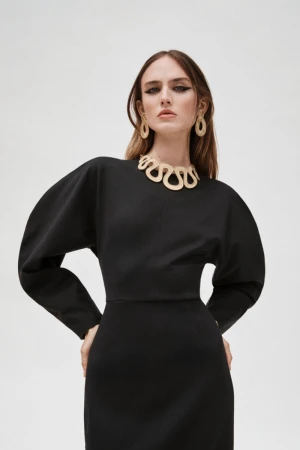 Studio shot of a model in a black midi dress with statement gold jewelry, posing against a plain white backdrop, promoting fashion.