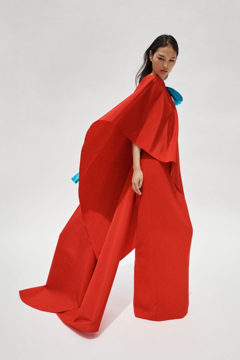 Elegant fashion shot featuring an Asian woman in a red dress with a dramatic cape, set against a clean, bright studio backdrop.