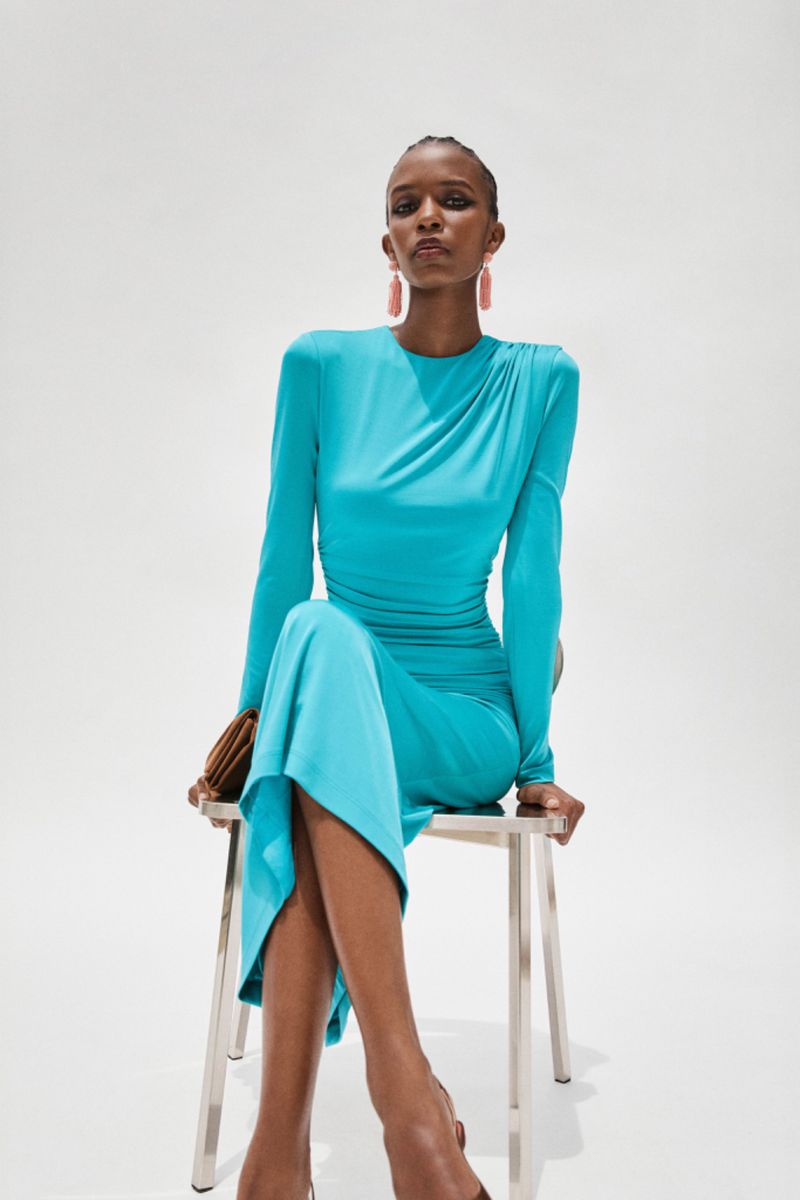A slim African-American woman with short hair sits on a stool wearing a turquoise dress and large earrings in a fashion studio shot.