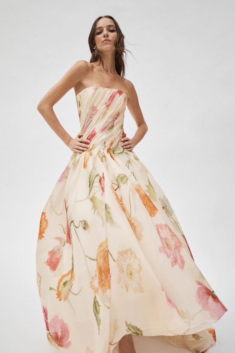 A woman poses in a strapless, floral-print gown with a flowing skirt against a white backdrop, showcasing a blend of fashion and art.