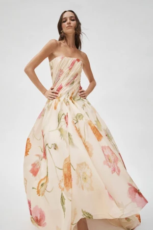 A woman poses in a strapless, floral-print gown with a flowing skirt against a white backdrop, showcasing a blend of fashion and art.