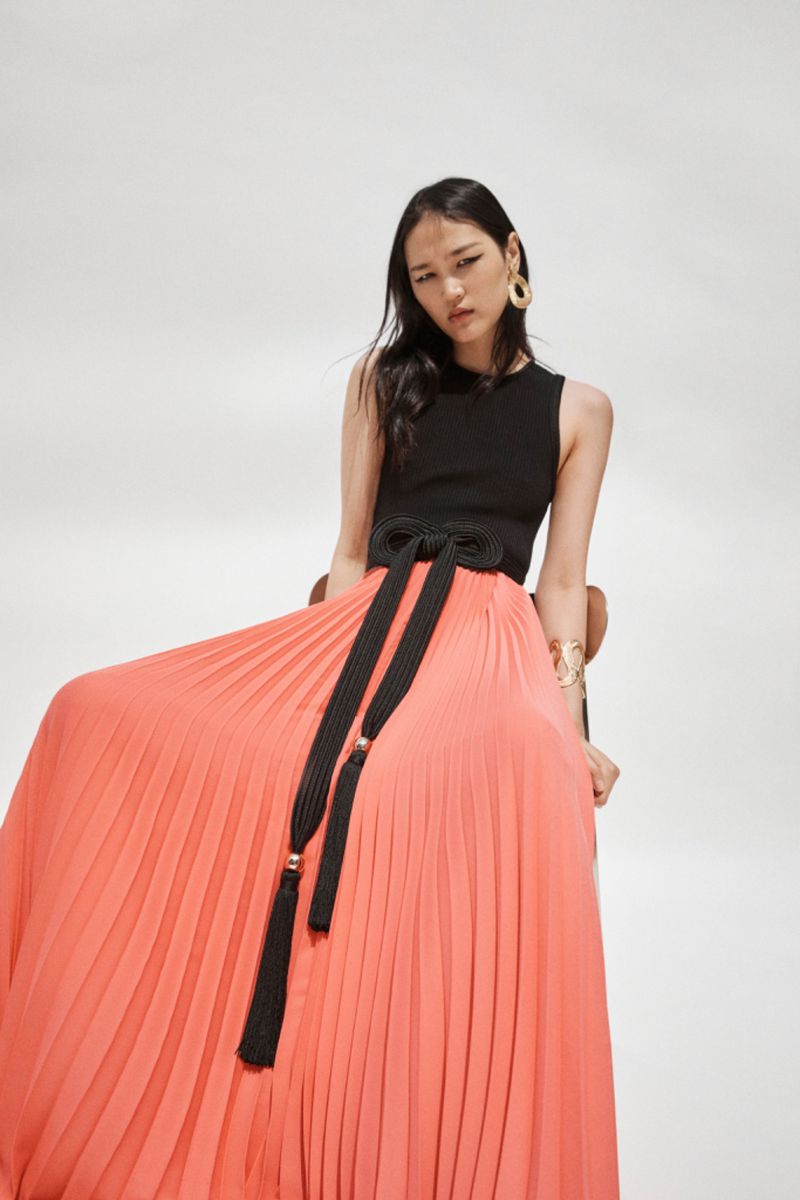 Fashion shoot of an Asian woman in a black top and pleated coral maxi skirt with a black rope belt and gold accessories, against white backdrop.