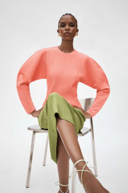 An African-American woman is seated on a chair in a studio setting, modeling a pleated coral top and green skirt, accessorized with white heels.