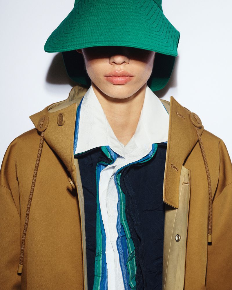 Woman poses indoors wearing a green sun hat, brown jacket, and a layered white and striped shirt against a white backdrop.