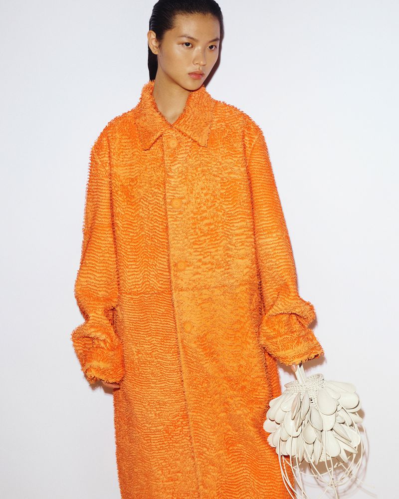 Fashion photo of an Asian woman wearing a textured orange coat with a unique handbag against a white backdrop, shot in studio lighting.