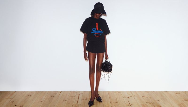 Woman in a black t-shirt and skirt with a Loewe logo, standing in a studio with a wood floor and white background.