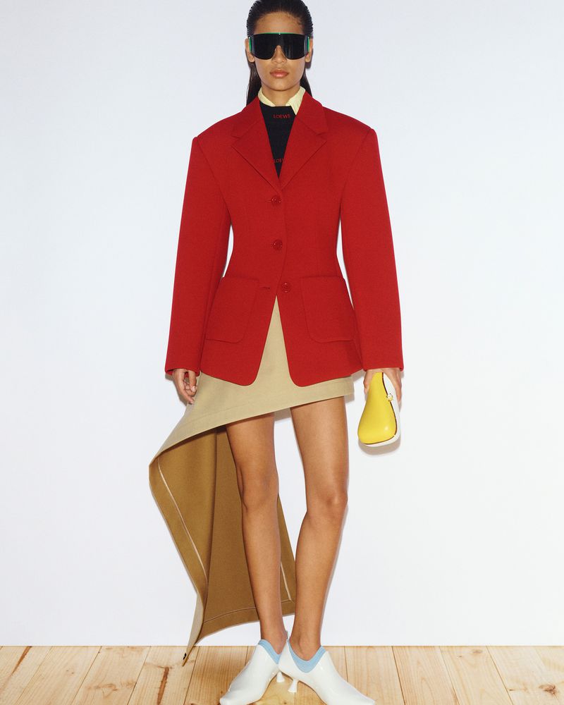 Fashion editorial shot of a Black female model in red blazer, khaki skirt, and white shoes against a white backdrop in studio lighting.