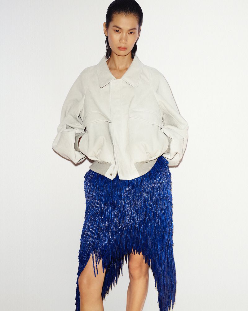 A stylish Asian woman poses in a studio wearing a light beige jacket and a striking blue fringe skirt, captured in a medium shot.
