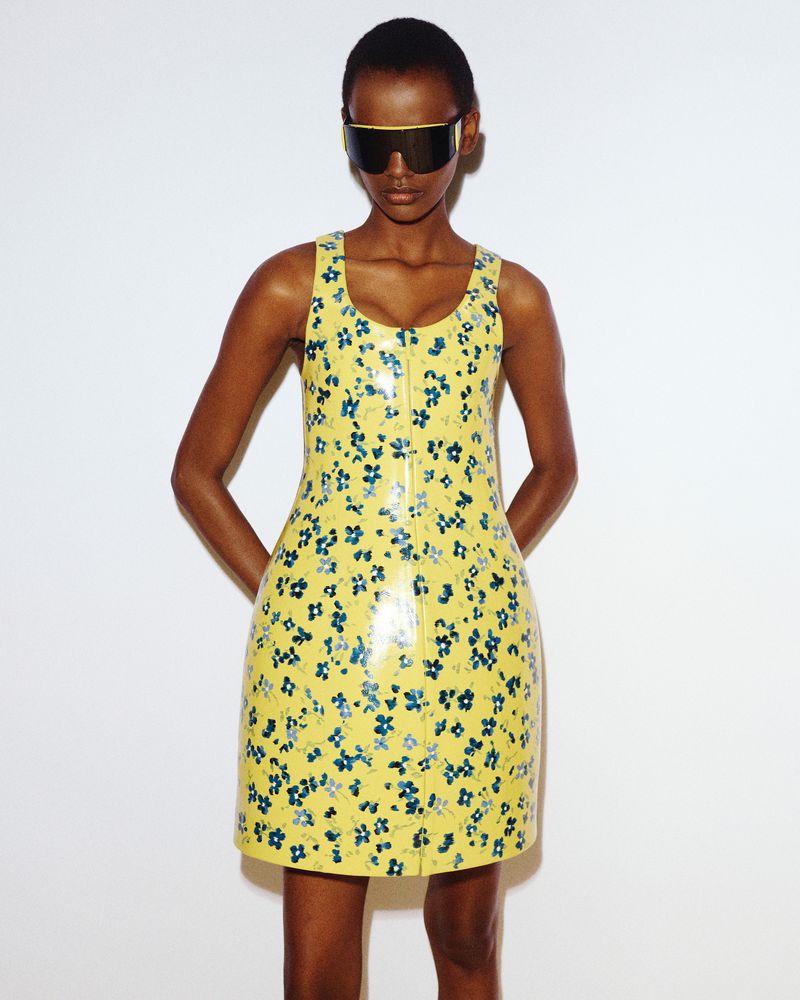 Black female model wearing a yellow floral sleeveless mini dress and oversized visor sunglasses against a white background.