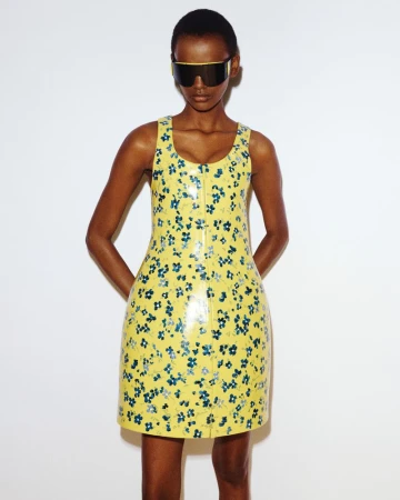 Black female model wearing a yellow floral sleeveless mini dress and oversized visor sunglasses against a white background.