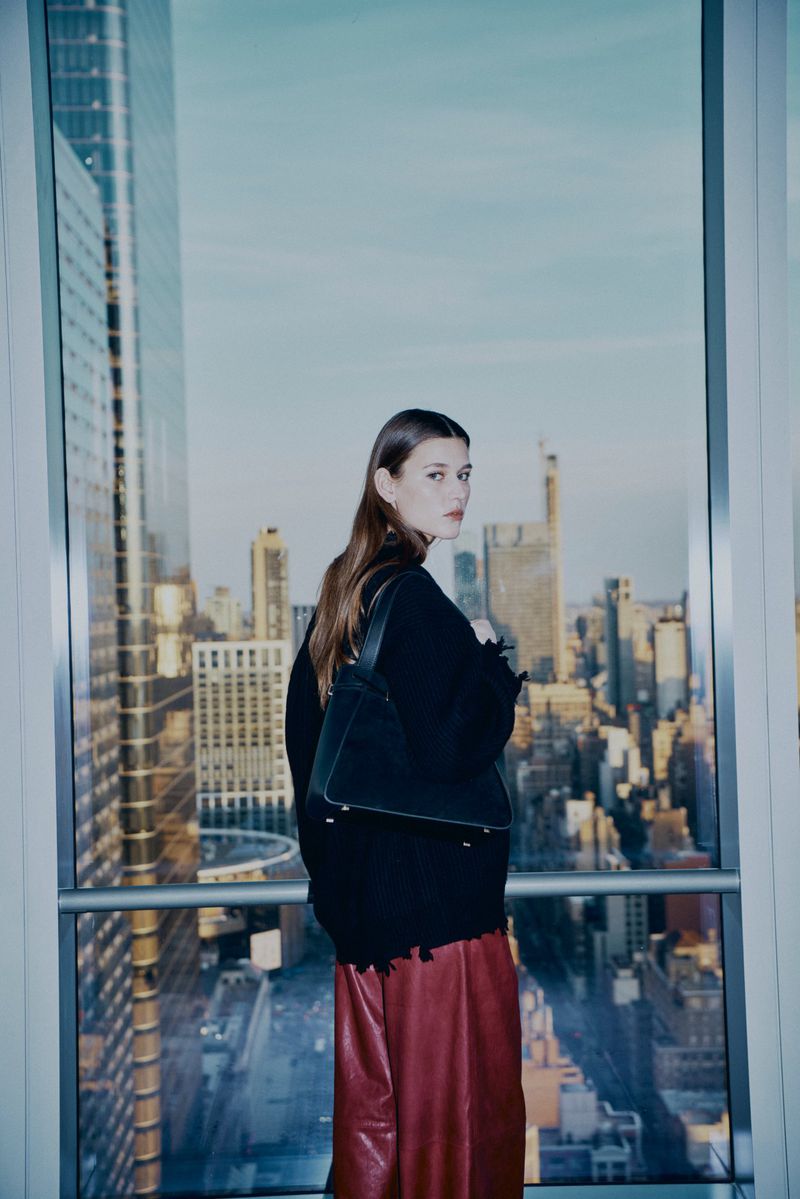 Woman with long brown hair wearing a black top and red pants with a handbag poses near a window overlooking a city skyline.
