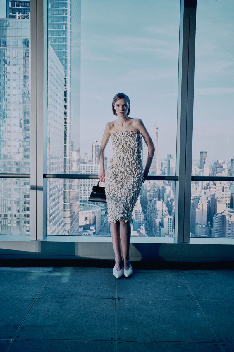 Fashionable woman in a strapless dress poses with a purse by a window overlooking a city skyline, bathed in cool, artificial light.
