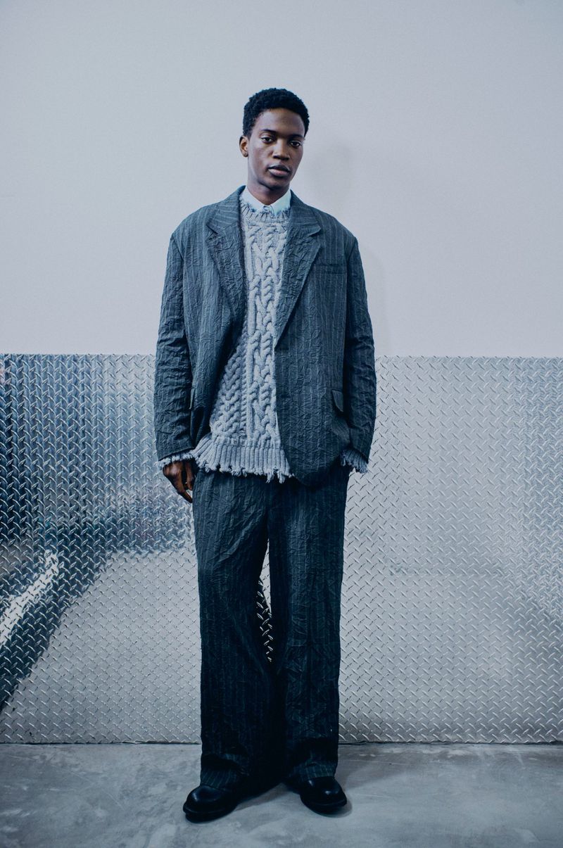 Full-length shot of an African-American male model wearing a striped grey blazer, pants, and a cable-knit sweater against a silver background.