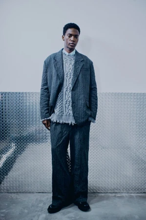 Full-length shot of an African-American male model wearing a striped grey blazer, pants, and a cable-knit sweater against a silver background.