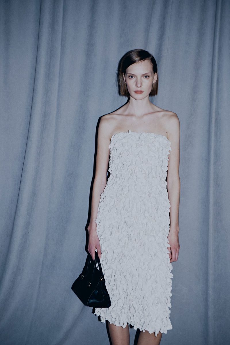 A woman in a white strapless dress and a black triangular purse poses in front of a grey curtain, showcasing a fashion editorial shot.