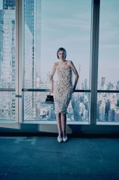 Model wearing a strapless textured white dress holding a black handbag in front of a floor-to-ceiling city skyline window