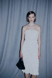 Model with a short bob wearing a strapless white textured dress and holding a black handbag in front of draped blue-grey curtains.