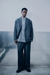 Model Limamu Mbaye wearing an oversized charcoal grey suit and cable-knit sweater for Simkhai against a diamond plate wall.