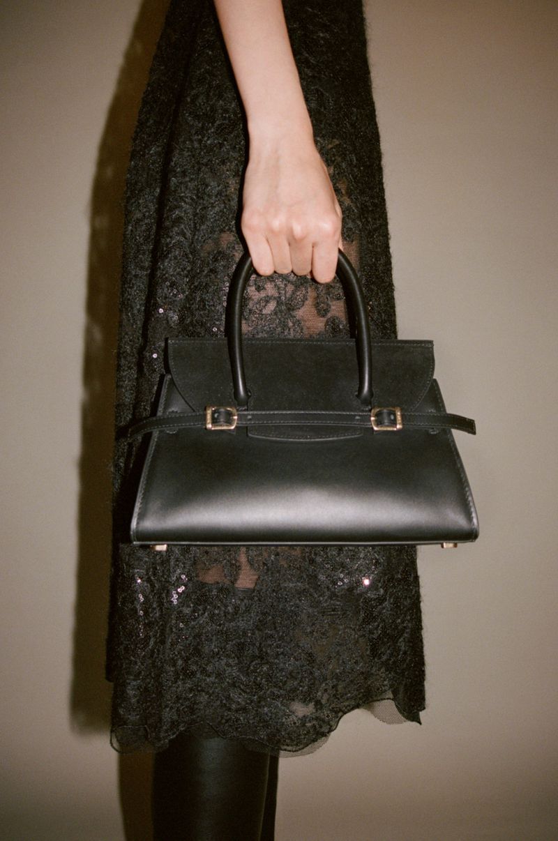 A black leather top-handle handbag with gold hardware is held by a woman with a sequined lace dress in a studio setting.