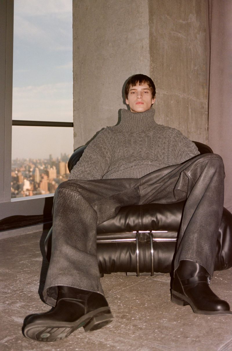 Man poses in grey cable-knit turtleneck, grey pants, and black boots in a modern interior with a cityscape view from the window.