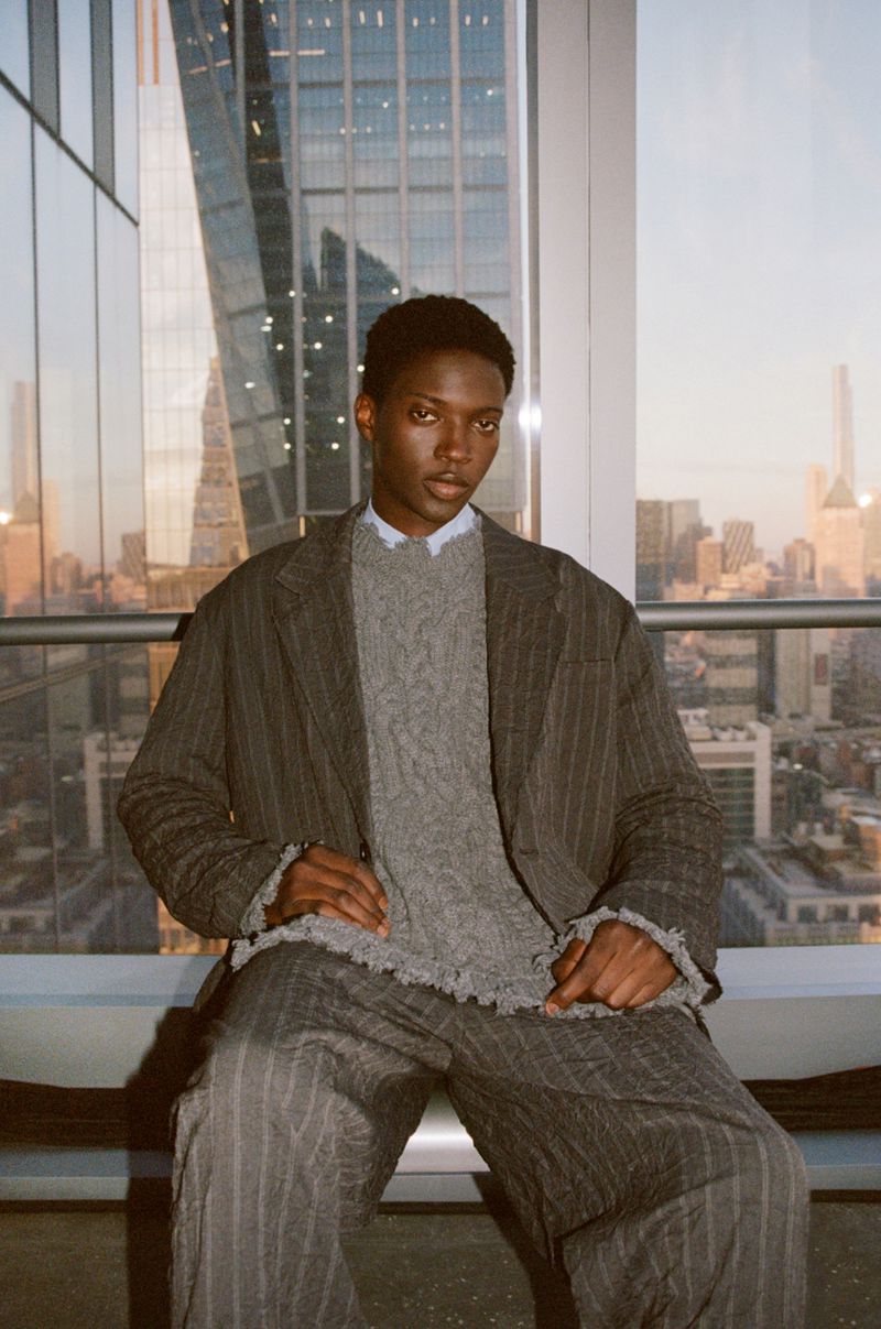 An African American man sits in a striped suit and grey sweater with a city view behind him, shot in portrait orientation.