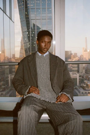 An African American man sits in a striped suit and grey sweater with a city view behind him, shot in portrait orientation.